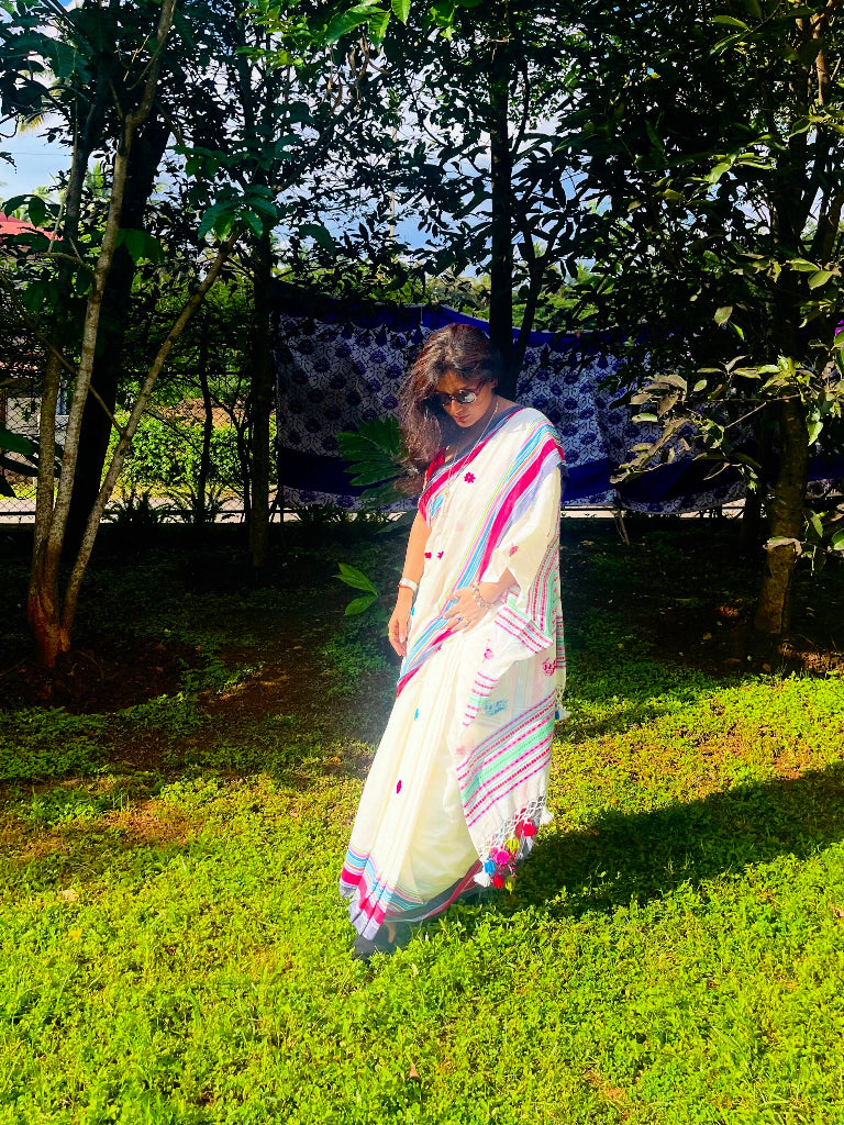 Woman in a white saree with colorful borders standing on grass with trees in the background